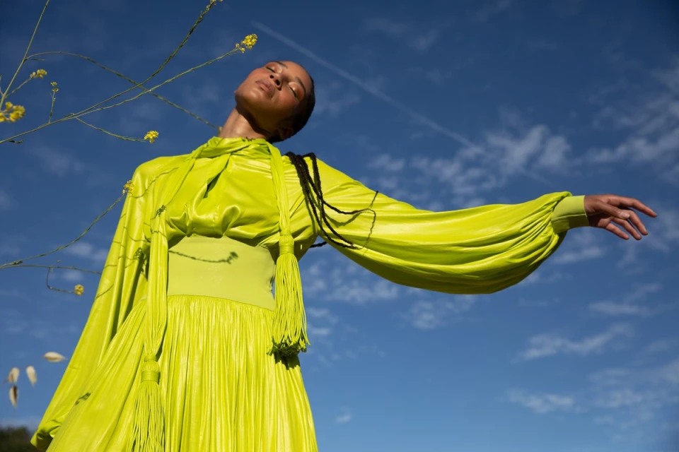 Adult female in bright green pleated gown posing with arms outstretched against a bright blue, slightly cloudy sky in an outdoor setting.