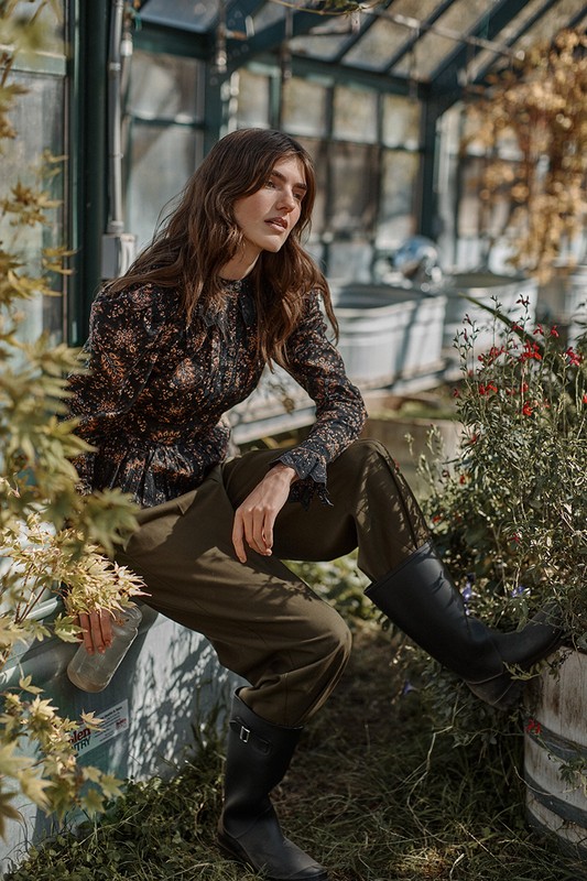 A woman in a long-sleeved floral top and olive pants sits by a greenhouse with plants and flowers around her, holding a bottle.