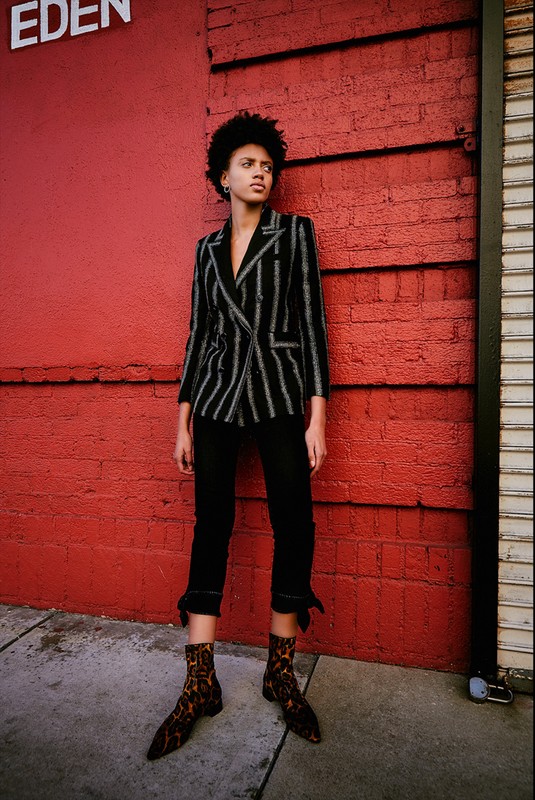 Adult woman in a striped blazer and black pants poses against a red brick wall on a city street in an eye-level medium shot.