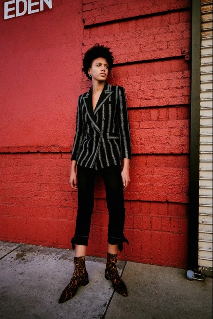 Adult woman in a striped blazer and black pants poses against a red brick wall on a city street in an eye-level medium shot.
