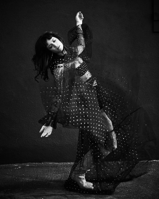 Black and white portrait of an adult woman in a polka-dot dress, dancing against a textured wall, striking an elegant pose.