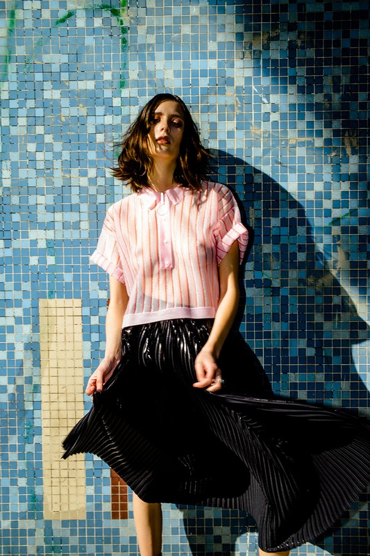 Young woman posing in a sheer pink top and black pleated skirt against a blue tiled wall in an eye-level, medium shot.