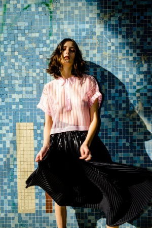 Young woman posing in a sheer pink top and black pleated skirt against a blue tiled wall in an eye-level, medium shot.