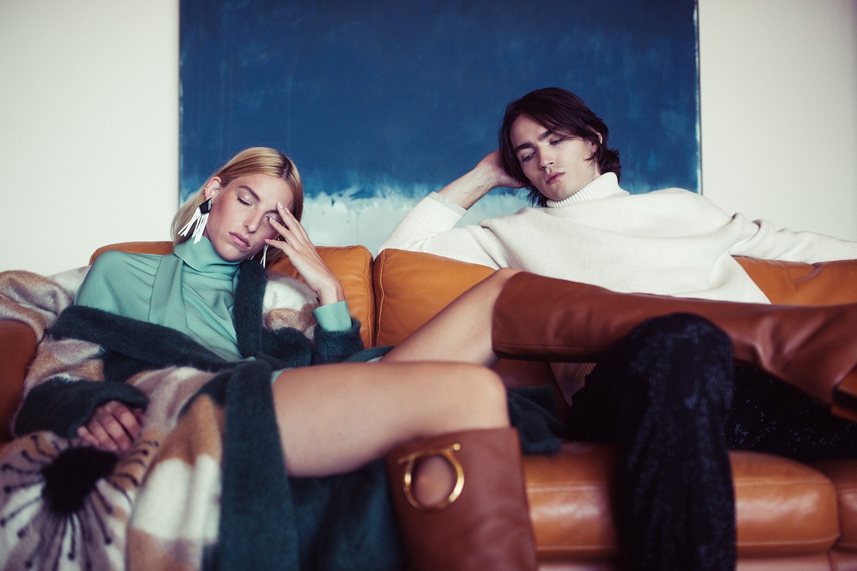 Two fashion models lounge on an orange couch in a stylish indoor setting. One wears a green top and the other a cream turtleneck.