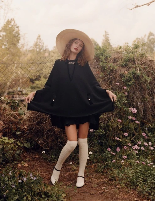 Adult woman wearing a large sunhat and dark poncho poses in a natural setting with flowers and foliage in a medium shot.