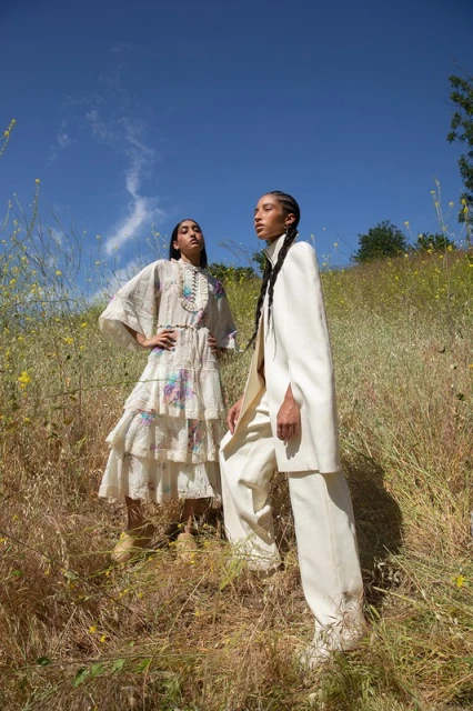 Two women pose in prairie grass under a blue sky. One woman wears a white dress with colorful embroidery, the other a white pantsuit.