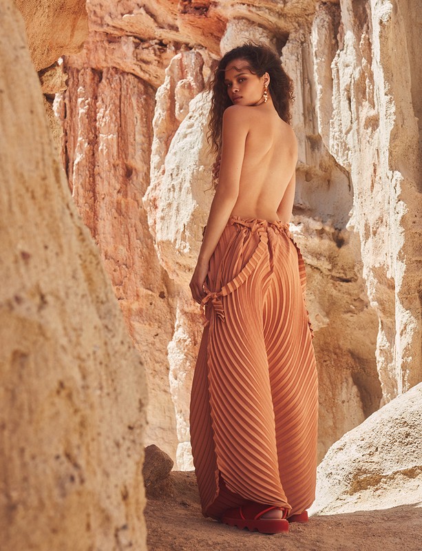 An adult female posing in a desert environment, showcasing wide-leg pleated pants and bare back in a fashion style editorial.