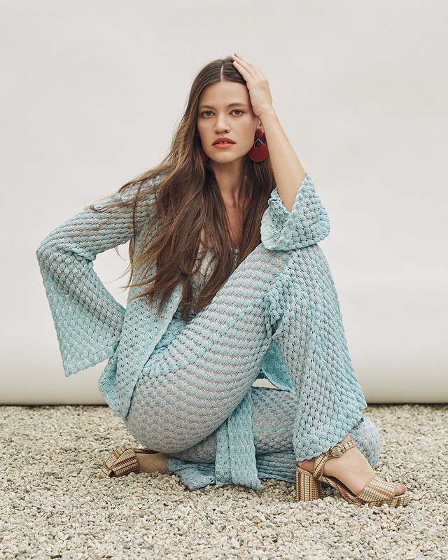 Brunette woman with hand on head poses in textured aqua top and pants with platform heels in a neutral, light-filled studio.