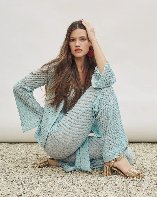 Brunette woman with hand on head poses in textured aqua top and pants with platform heels in a neutral, light-filled studio.
