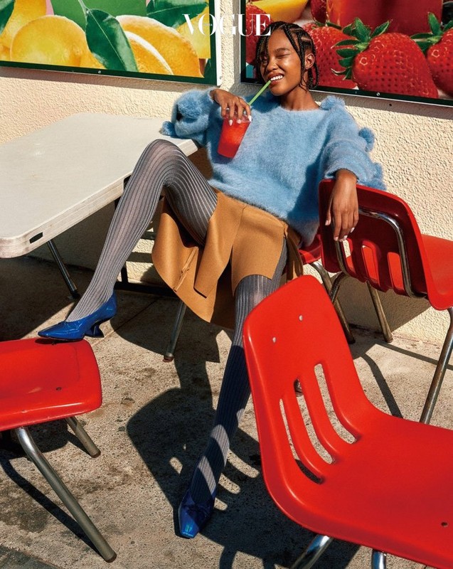 Woman in blue sweater, tan skirt, and gray tights sits at a table with a drink. Vogue magazine cover in background. Fashion editorial.
