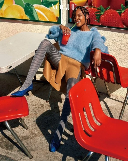 Woman in blue sweater, tan skirt, and gray tights sits at a table with a drink. Vogue magazine cover in background. Fashion editorial.
