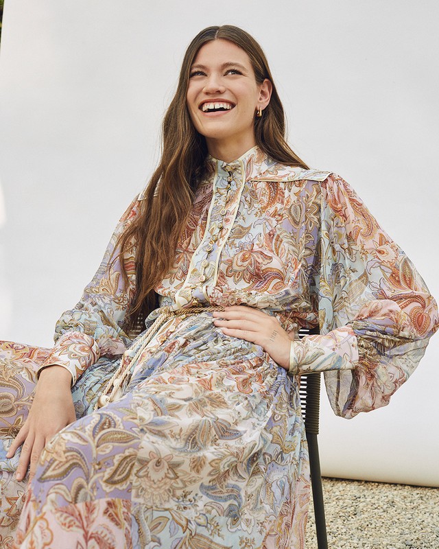 A woman with long brown hair smiles while wearing a floral patterned dress in a light and airy setting against a neutral backdrop.