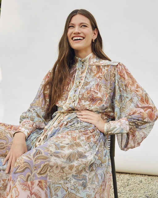 A woman with long brown hair smiles while wearing a floral patterned dress in a light and airy setting against a neutral backdrop.