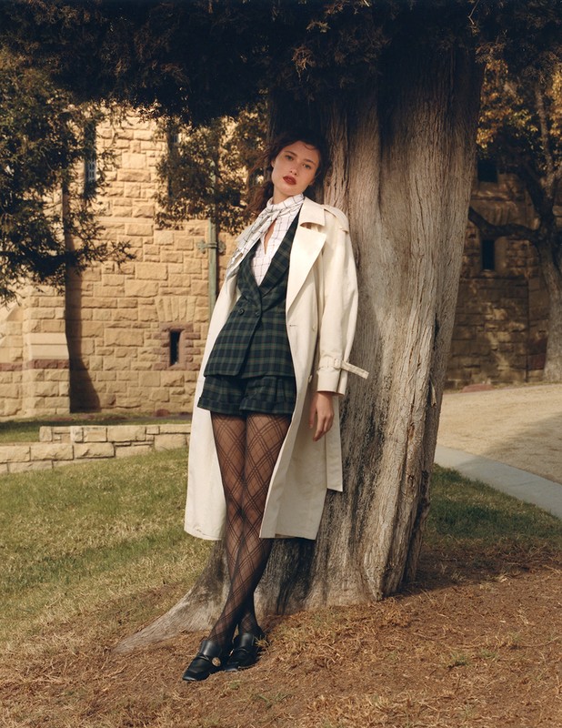 A young adult female model poses leaning against a tree wearing a beige trench coat, plaid mini skirt suit and black tights in daylight.