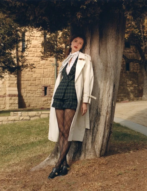 A young adult female model poses leaning against a tree wearing a beige trench coat, plaid mini skirt suit and black tights in daylight.