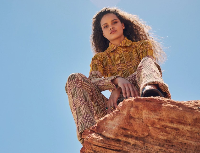 Young woman with curly hair in a plaid suit sits on a rock formation against a bright blue sky in a low-angle fashion-style photograph.