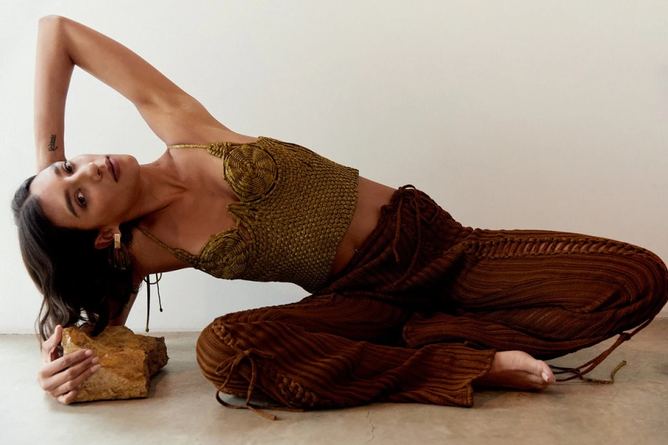 Woman poses in a gold knit halter top and brown crinkle pants with a rock, styled in an eye-level indoor fashion shot.