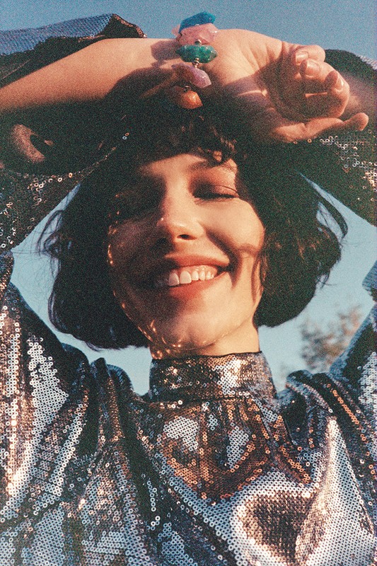 Portrait of smiling woman with short dark hair in shimmery silver top, raising hands with a jeweled ring on fingers outdoors.