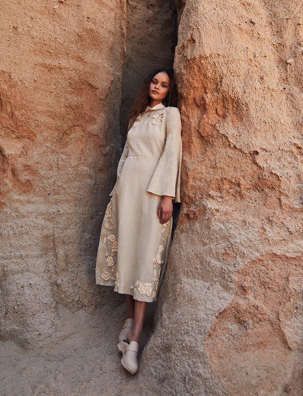 A woman in a beige dress stands between rocky cliffs, showcasing a blend of fashion and nature in a neutral-toned outdoor setting.