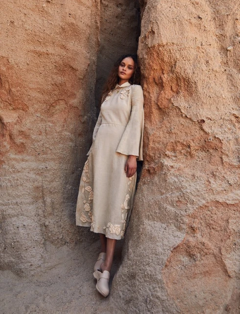 A woman in a beige dress stands between rocky cliffs, showcasing a blend of fashion and nature in a neutral-toned outdoor setting.
