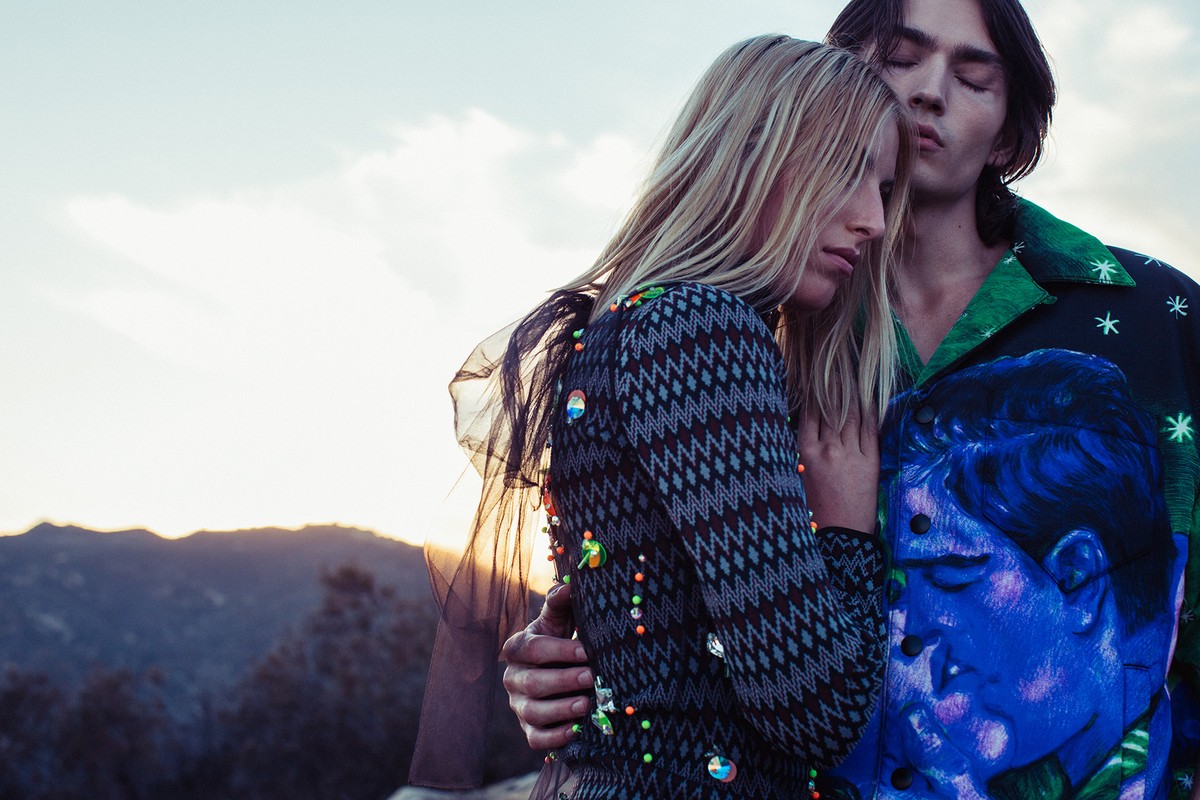 A stylish couple poses outdoors at sunset: a blonde woman in patterned dress and a man in graphic shirt, captured in a close-up fashion shot.