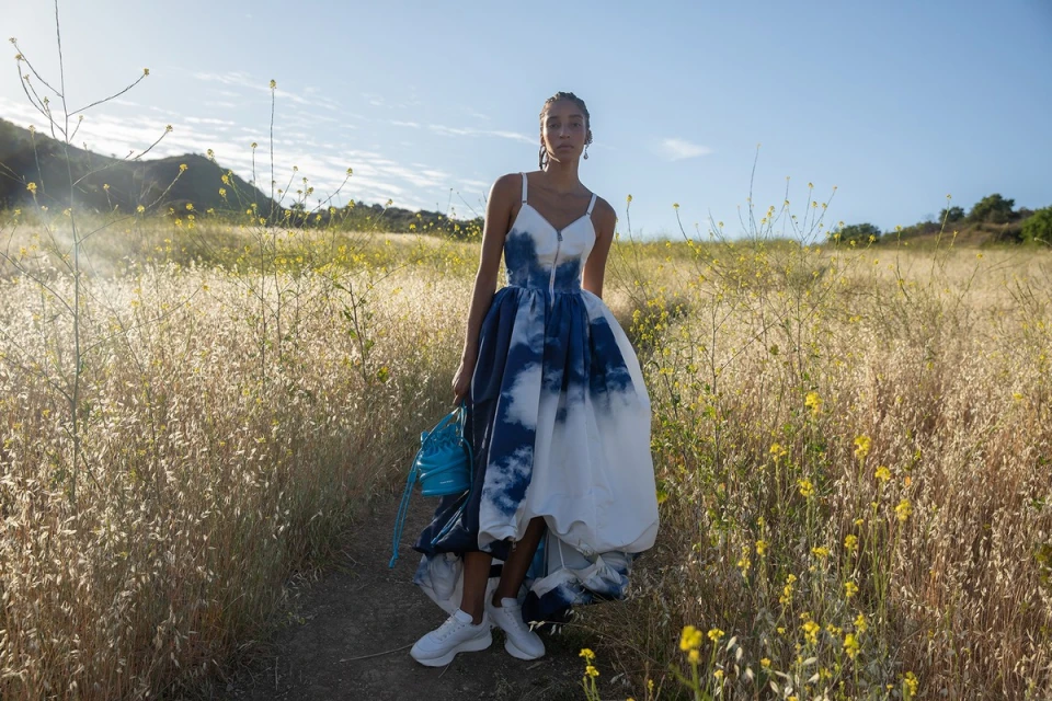 Woman in flowing blue-and-white gown stands on grassy path with blue bag and white sneakers in outdoor fashion shot in sunny meadow.