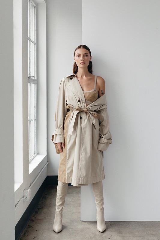 A stylish adult woman poses in a beige trench coat and matching boots near a window in an indoor setting against a white wall.
