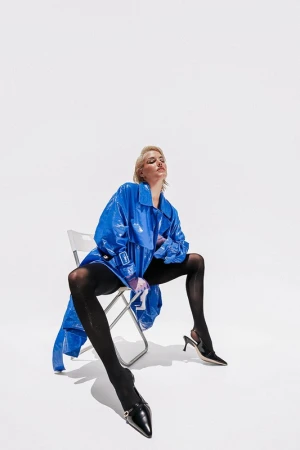 Model with blonde hair wearing a glossy blue coat and black tights seated on a folding chair against white