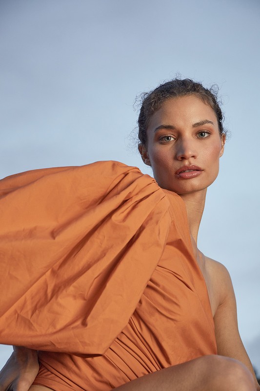 Portrait of a woman with curly hair and one-shoulder orange dress against light blue sky. Fashion, beauty and outdoor concept.