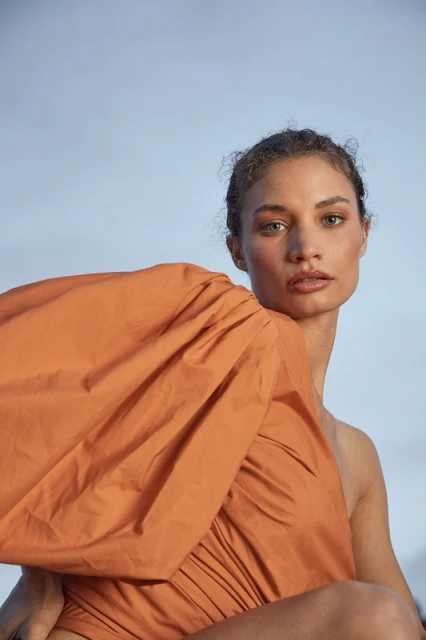 Portrait of a woman with curly hair and one-shoulder orange dress against light blue sky. Fashion, beauty and outdoor concept.