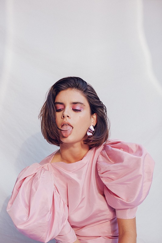 Portrait of a young brunette woman in a pink dress with puffy sleeves, sticking her tongue out, posing in a bright photo studio.
