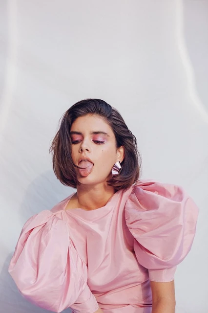 Portrait of a young brunette woman in a pink dress with puffy sleeves, sticking her tongue out, posing in a bright photo studio.
