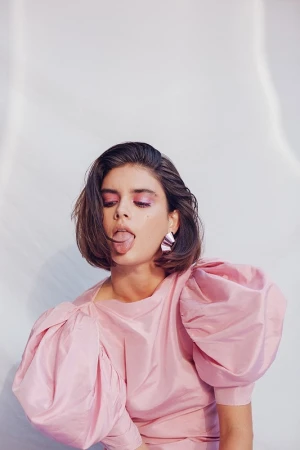 Portrait of a young brunette woman in a pink dress with puffy sleeves, sticking her tongue out, posing in a bright photo studio.