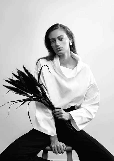 Elegant portrait of a young woman with black hair, wearing white top and black pants while holding dark feathery stems, in studio lighting.