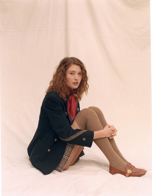 Young woman with auburn curly hair in a dark blazer, reddish scarf, plaid skirt, knee socks, and loafers posing on a white backdrop.