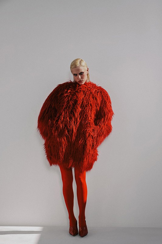 Woman with fair hair poses in a red faux fur coat, red tights, and red high heels against a plain background in a studio setting.