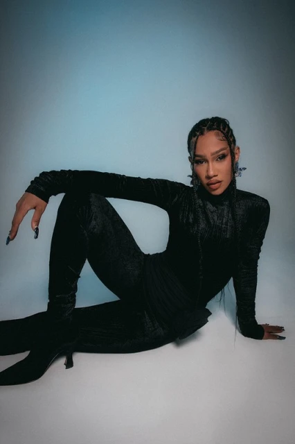 Portrait of a stylish adult female model wearing a black velvet jumpsuit and heels while posing against a blue gradient backdrop.
