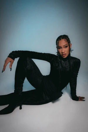 Portrait of a stylish adult female model wearing a black velvet jumpsuit and heels while posing against a blue gradient backdrop.