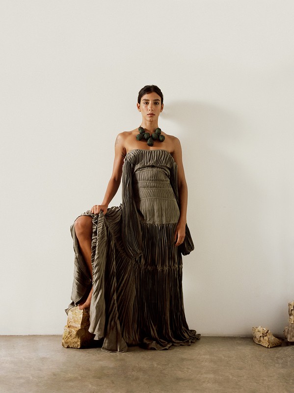 A woman in an olive green gown and necklace poses on a rock in front of a blank wall in a studio setting. Fashion editorial.