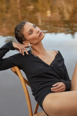 Woman with short dark hair in a black long-sleeve top sitting on a wooden chair outdoors near water.