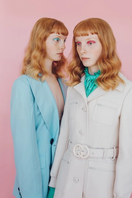 Two red-haired women pose in matching outerwear against a pink background, one in a turquoise blazer, the other in a white coat.