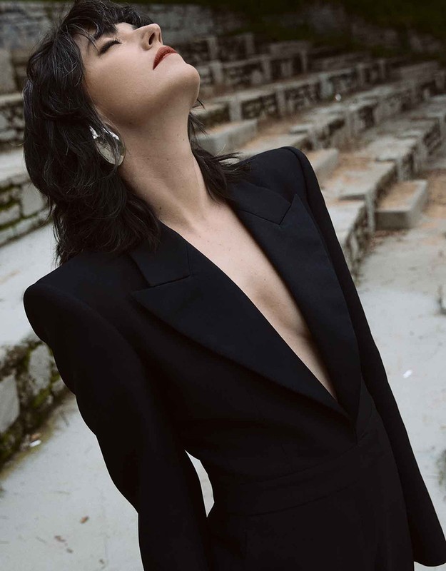 Woman in a black blazer and large silver earrings looks up outdoors. The stone steps of an amphitheater are in the background.