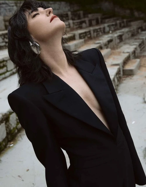 Woman in a black blazer and large silver earrings looks up outdoors. The stone steps of an amphitheater are in the background.