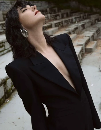 Woman in a black blazer and large silver earrings looks up outdoors. The stone steps of an amphitheater are in the background.