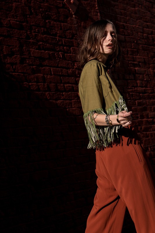Woman in an olive green fringed top and rust orange trousers posing against a dark brick wall.