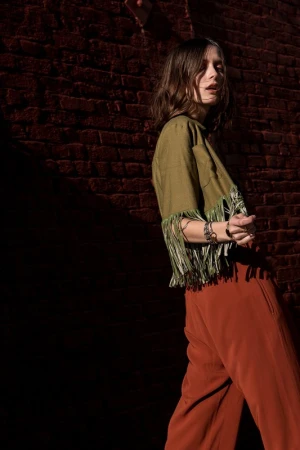 Woman in an olive green fringed top and rust orange trousers posing against a dark brick wall.