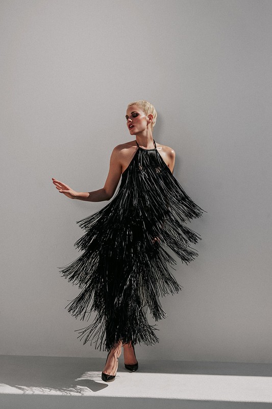 Adult female in black fringe dress and high heels poses against gray backdrop in studio lighting with soft shadows, fashion photography.