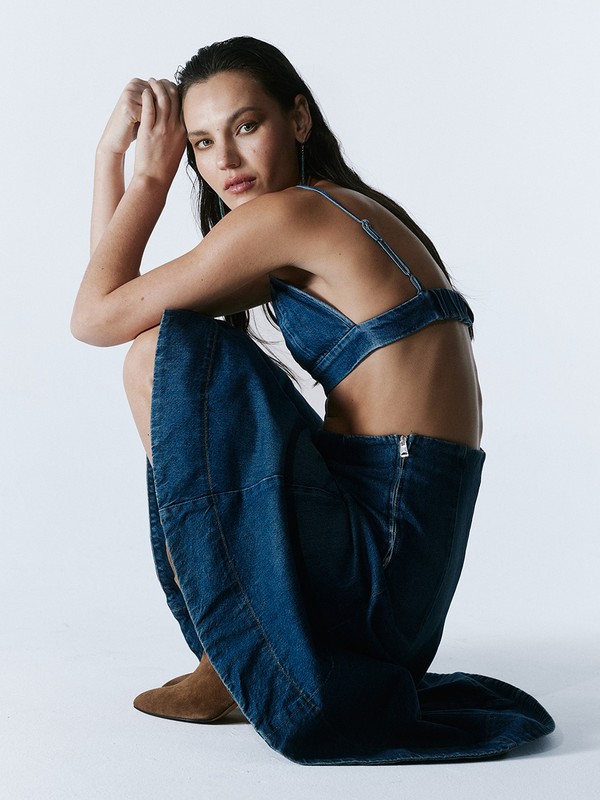 Brunette woman in a denim crop top and skirt crouches in a photo studio, looking at the camera with a neutral expression.