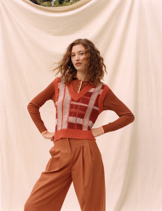 A young woman poses in an outfit with a brown color palette, wearing a knit sweater vest and culottes against a draped backdrop.