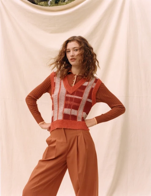 A young woman poses in an outfit with a brown color palette, wearing a knit sweater vest and culottes against a draped backdrop.